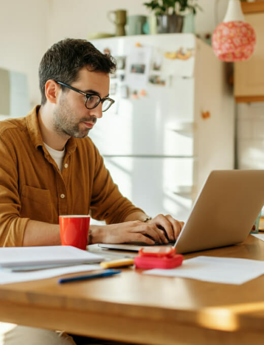 A developer typing on a laptop at a kitchen table beside notes and a coffee mug